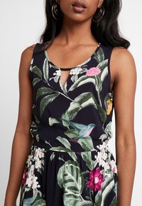 Ärmlös blommig topp i marinblå som har stora gröna blad, rosa och vita blommor, en liten nyckelhålsdetalj vid halsen och en avslappnad passform.