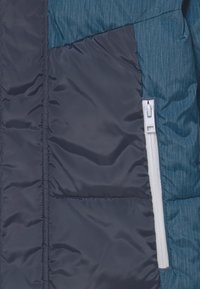 Close-up van een blauwe gestikte jas met een witte verticale rits en een contrasterend donker stoffen paneel naast de rits aan de voorkant.