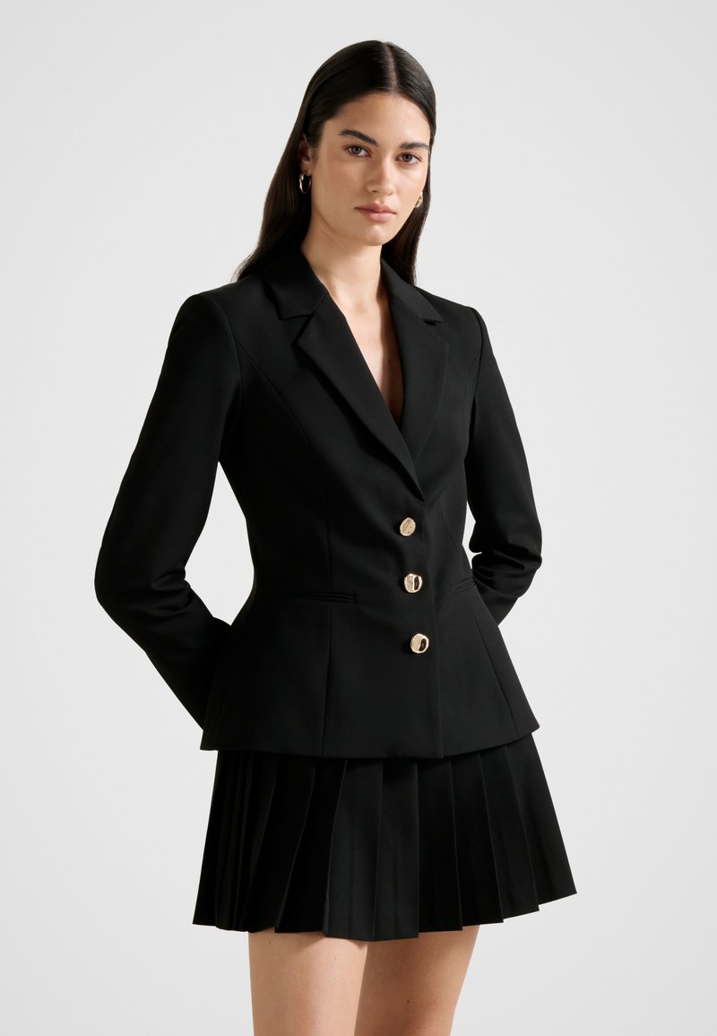 Kvinde med langt mørkt hår iført en figursyet sort blazer med guldknapper og en sort plisseret nederdel, stående foran en ensfarvet baggrund.