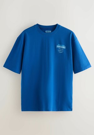 Zila kokvilnas t-kreklus ar īsām piedurknēm, ar balta logo drukājumu kreisajā krūtīs. Gluda tekstūra un brīva piegriezuma forma.