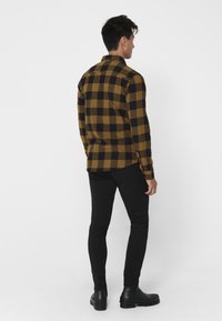 Heren geel en zwart geblokt flanellen overhemd met lange mouwen, gecombineerd met zwarte slim-fit broeken en zwarte laarzen, gezien vanaf de achterkant.