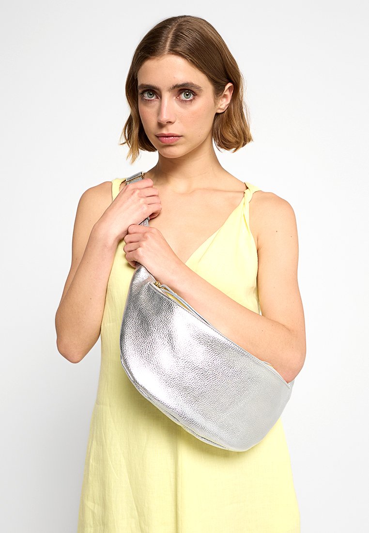 Eine Frau mit kurzen braunen Haaren, die ein ärmelloses gelbes Kleid trägt und eine große, silberne, strukturiertes Schultertasche vor einem weißen Hintergrund hält.