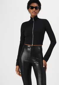 Giacca nera cropped con zip e maniche lunghe, abbinata a leggings neri lucidi a vita alta con un piccolo emblema vicino all'elastico.
