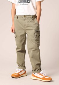 Khaki cargobroek met twee zijzakken met rits, een knoopsluiting en omslagen aan de broekspijpen. Gepaard met oranje en witte sneakers met textuurelementen.