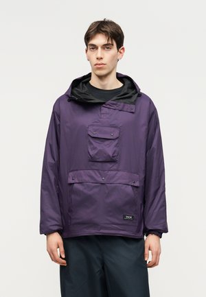 Young man wearing a loose purple hooded jacket with front pockets and black pants, standing against a plain light background.