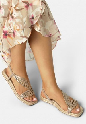 Pies con sandalias doradas y brillantes con tiras en forma de hoja, combinadas con una falda beige con flores rosas y naranjas.