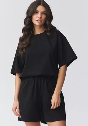 Kobieta z długimi falującymi włosami, ubrana w luźną czarną koszulkę z krótkim rękawem i dopasowane czarne szorty, na jasnoszarym tle.