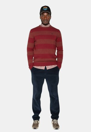 Pull rayé bordeaux, chemise à motifs floraux en dessous, pantalon en velours côtelé navy, baskets beiges et blanches, casquette noire avec un logo de montagne.