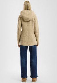 Beige villasegu segu kombineeritud mantel kapuutsiga, millel on tekstuuriga muster ja sobiv disain, koos tumehallide laia säärega teksade ja pruunide saapatega.