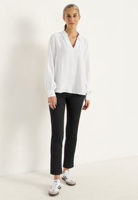 Vit långärmad blus med V-halsringning och indragna manschetter, i kombination med svarta åtsittande byxor och vita sneakers med svarta och blåa detaljer.