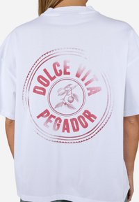T-shirt oversized blanche avec un imprimé circulaire rouge comportant "DOLCE VITA PEGADOR" et un graphique de feuilles et de cerises. Tissu en coton lisse.
