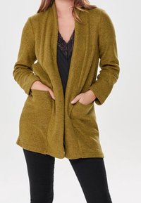 Olivgrön, texturerad cardigan med öppen front och två sidofickor, matchad med en svart spetsblus och svarta byxor; ärmar i medellängd.