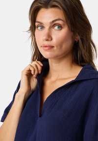 Chemise bleu marine avec col, en tissu texturé, manches trois-quarts et un détail de logo discret sur le devant. Main saisissant le col.