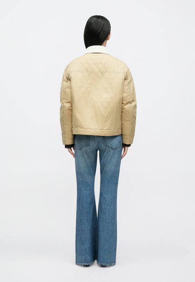 Beežikas paks jakk valge karusnahast kraega, vaadates tagant. Ühitatud laia säärega siniste teksade ja tumeda aluspaelaga.