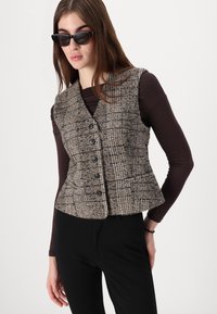 Brūns šaurs vests ar rūtainu rakstu, kam ir seši melni pogas, apvienots ar brūnu garām piedurknēm krekli un melnām biksēm. Gluda tekstūra un pielāgota forma.