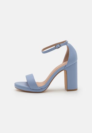 Sandale à talon haut bleu clair avec un talon épais, orteils ouverts, une bride de cheville et une texture en cuir lisse. Semelle plate avec un rembourrage minimal.