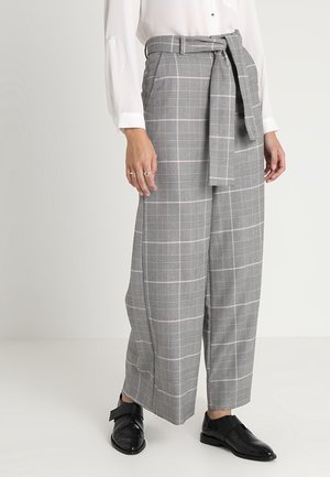 Femme portant un pantalon large à carreaux gris avec ceinture, chemise blanche boutonnée et chaussures en cuir noires sur un fond clair uni.