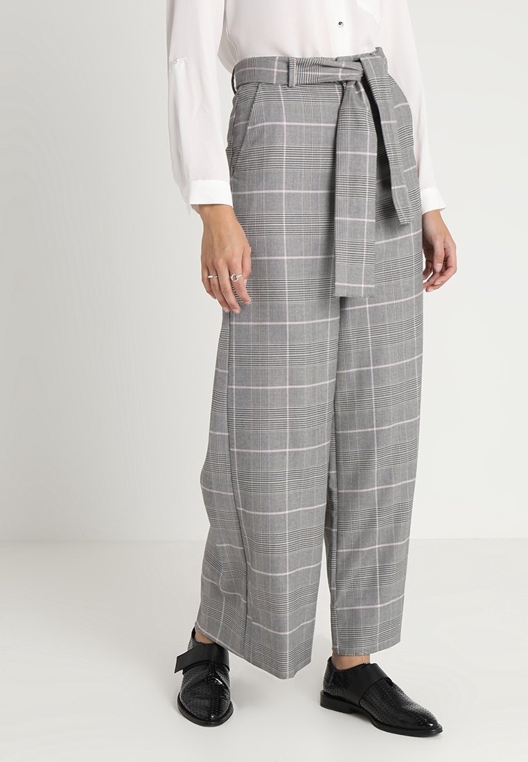 Femme portant un pantalon large à carreaux gris avec ceinture, chemise blanche boutonnée et chaussures en cuir noires sur un fond clair uni.