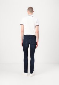 Vit polo skjorta med röda detaljer, kombinerad med mörkblå tajta jeans. Modellen har på sig vita sneakers och ses bakifrån.