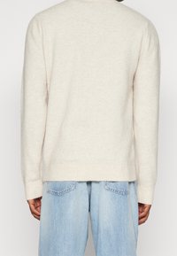 Ljusbeige ullblandad tröja med ribbat nederkant och ärmslut, som har en avslappnad passform och mjuk textur. Bärs tillsammans med ljusblå denimjeans.