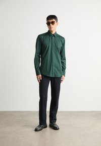 Camisa verde de botones con cuello, de manga larga y textura suave, combinada con pantalones oscuros y zapatos negros. Se han añadido gafas de sol marrones.