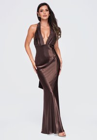 Femme aux longs cheveux foncés portant une robe en satin marron brillante sans manches avec un décolleté en V profond et des talons ouverts beige sur un fond clair uni.