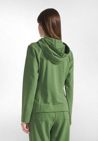 Sudadera con capucha verde hecha de tela suave, con un ajuste holgado, mangas largas y capucha con cordón. Textura suave y detalles mínimos.