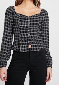 Haut à manches longues noir et blanc à carreaux avec un décolleté carré et une fermeture à boutons, présentant un style peplum au niveau de la taille.