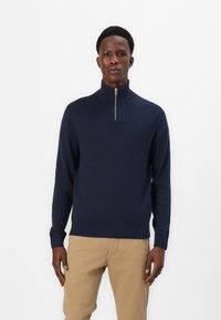 Marineblauer Strickpullover mit hohem Stehkragen und einem Viertelreißverschluss. Rippenbündchen und -saum; glatte Textur mit minimalen Designdetails.