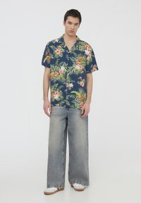 Camicia navy a maniche corte con motivo floreale e jeans a gamba larga azzurri; calzature bianche con suole in legno; sfondo neutro.