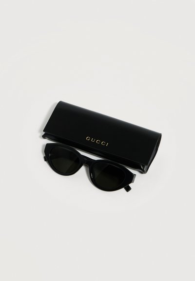 Μαύρα γυαλιά ηλίου με σχήμα cat-eye και σκοτεινούς φακούς, τοποθετημένα κάτω από μια μαύρη θήκη Gucci με χρυσά γράμματα σε λευκό φόντο.