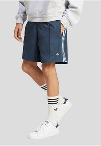 Jūrų mėlynumo sportiniai šortai su auksiniu logotipu, baltais šonais ir šoninėmis kišenėmis. Nešiojami su dryžuotomis kojinėmis ir baltais sportbačiais su jūrų mėlynais akcentais.
