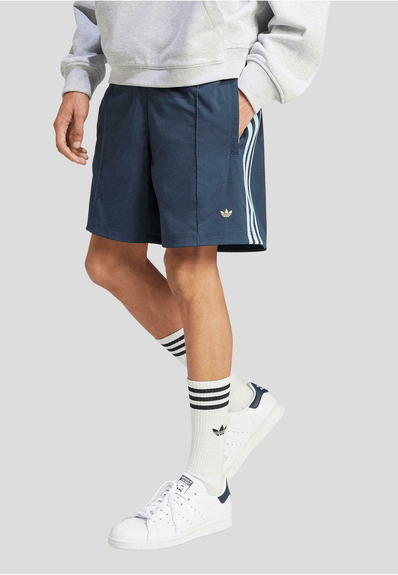 Shorts de sport bleu marine avec logo doré, rayures blanches sur les côtés et poches latérales. Portés avec des chaussettes rayées et des baskets blanches avec des accents bleu marine.