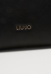 Détail d'un sac à main en cuir noir montrant le logo métallique doré LIU JO sur une surface texture.