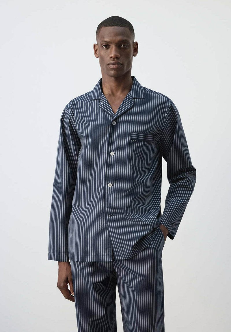 Homme portant une chemise bleu foncé à fines rayures et manches longues, ainsi qu'un pantalon assorti, debout devant un fond blanc uni.
