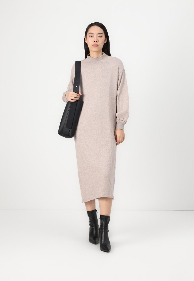 Sukienka swetrowa do kolan w jasnym beżu z bufkami i wysokim dekoltem. W komplecie z czarnymi botkami za kostkę oraz czarną torebką na ramię.