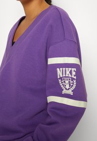 Personne portant un sweat-shirt Nike violet avec des rayures blanches et un logo indiquant "Nike 1972 Oregon" sur la manche, sur fond blanc.