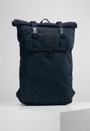 Sac à dos noir à enroulement supérieur avec rabat avant sécurisé par deux boucles et une poche zippée en dessous, posé sur une surface blanche.