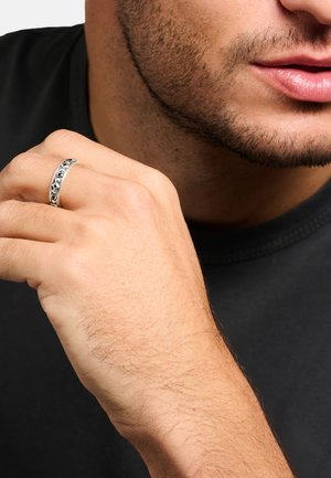 Nahaufnahme des unteren Gesichtsbereichs und der Hand eines Mannes, die einen silbernen Bandring mit schwarzen Sternmotiven an seinem Finger zeigt.