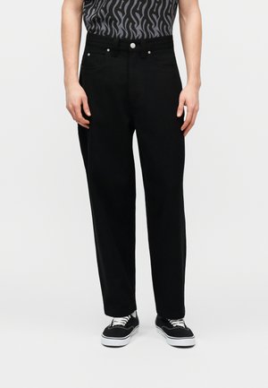 Hombre vestido con pantalones negros de pierna recta, zapatillas casuales blancas y negras, y una camisa gris con estampado, de pie contra un fondo liso.