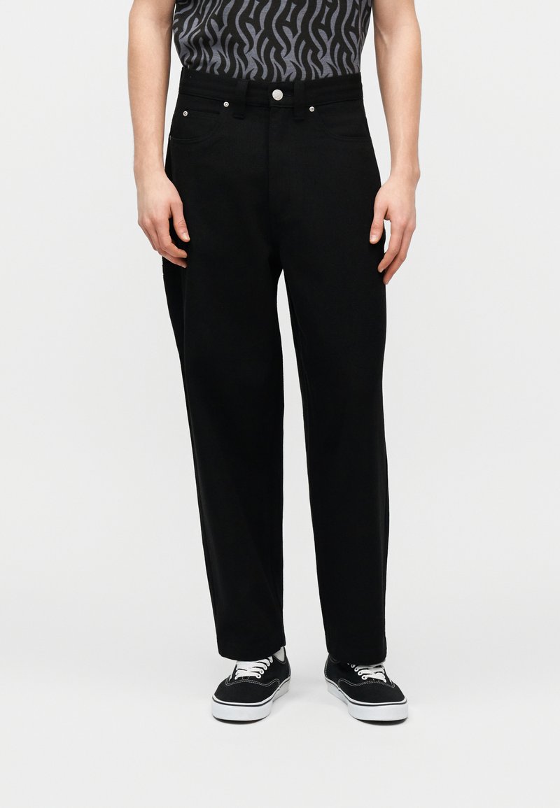 Άνδρας που φοράει μαύρο παντελόνι με ίσια γραμμή, μαύρα και λευκά casual αθλητικά παπούτσια και γκρι πουκάμισο με σχέδιο, στέκεται μπροστά σε μια απλή φόντο.