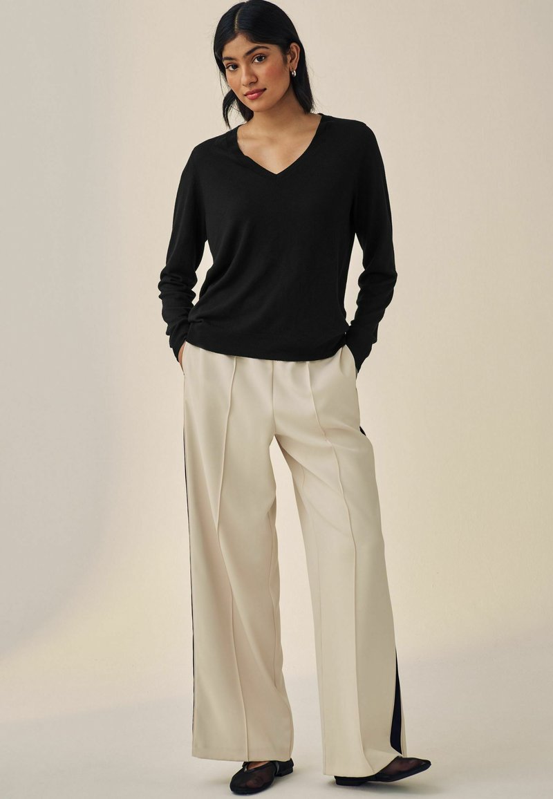 Un pull noir à col en V associé à un pantalon crème ample avec des fentes latérales et des accents noirs. Le modèle se tient avec les mains dans les poches.