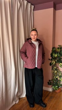 Νεαρή γυναίκα στέκεται μέσα σε εσωτερικό χώρο φορώντας μωβ καπιτονέ μπουφάν, λευκό πουκάμισο, φαρδιά μαύρη παντελόνια και μαύρα παπούτσια δίπλα σε ένα πράσινο γλαστράκι.