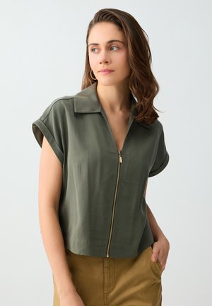 Jeune femme aux cheveux bruns mi-longs porte une chemise zippée manches courtes olive et un pantalon beige, regardant sur le côté avec la main dans la poche.