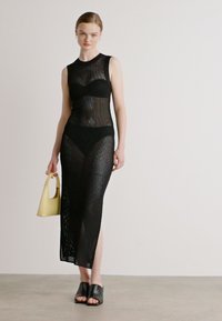 Rochie neagră din plasă cu un corset fixat și o fustă lungă cu slit. Purtați cu pantofi negri cu vârf deschis și ținând o geantă mică galbenă.