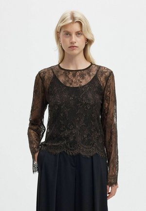 Un mannequin blonde porte un haut noir à manches longues en dentelle florale sur un caraco noir et un pantalon foncé, sur un fond blanc uni.