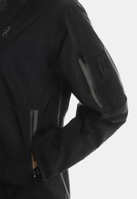 Czarna wodoodporna kurtka o gładkiej fakturze, z odblaskowymi akcentami, kieszeniami na zamek błyskawiczny oraz naszywką z logo na rękawie.
