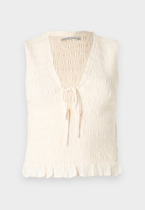 Haut sans manches crème avec un col en V, présentant une texture smockée et un détail de nouage à l'avant. Un ourlet à volants ajoute de la douceur au design.