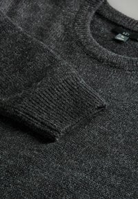 Ciemnoszary sweter z dzianiny z prążkowanymi mankietami i okrągłym dekoltem, leżący płasko z widoczną metką rozmiarową wewnątrz kołnierza.