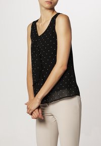 Blusa negra sin mangas con lunares blancos, escote en V y un diseño fluido y en capas en el frente. Textura de tela suave.